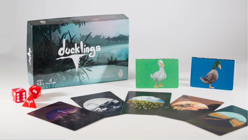 Ducklings tabletop game photographed with its illustrated box, duck cards, location cards and red dice.